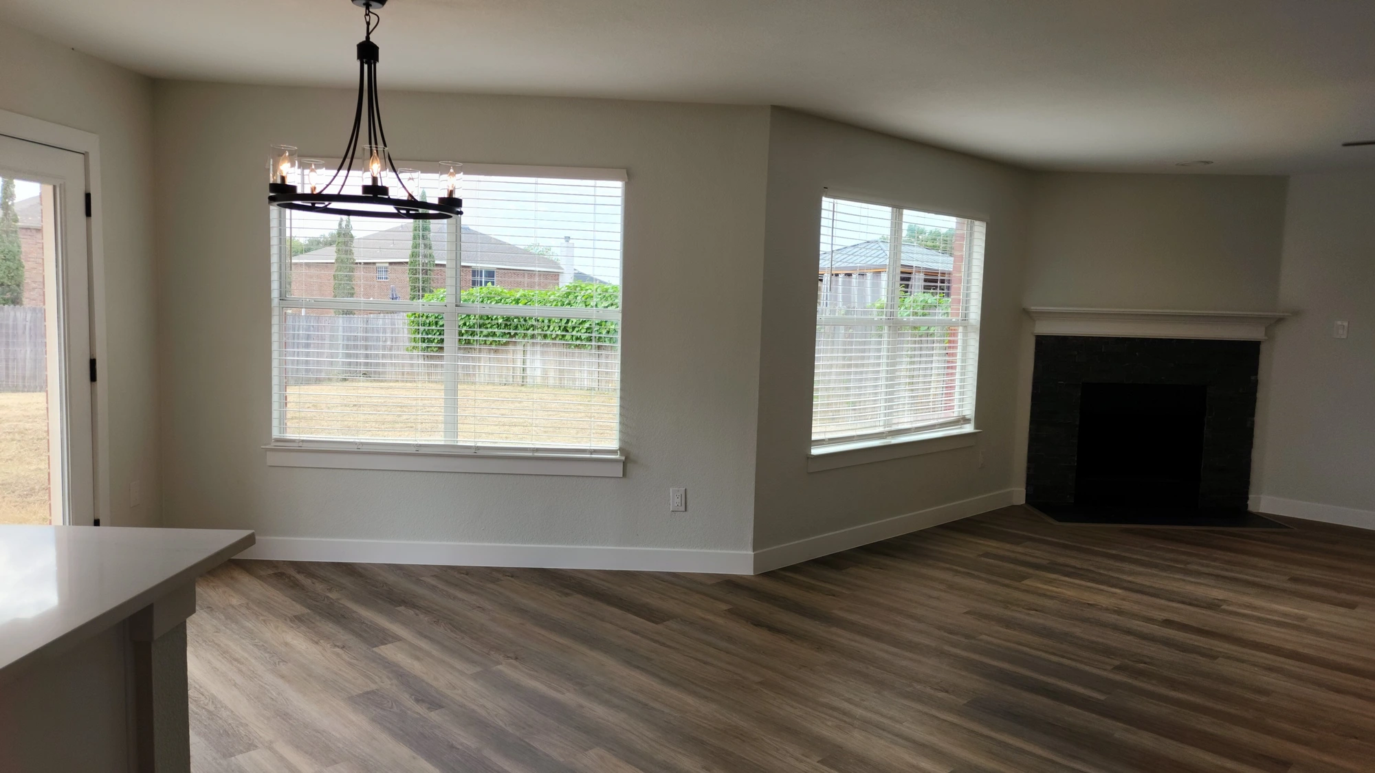After investor rehab: open living and dining room with medium-brown wood-look LVP flooring, light greige paint, wide white baseboards, updated corner fireplace surround with white mantel, black wagon-wheel chandelier, and white blinds