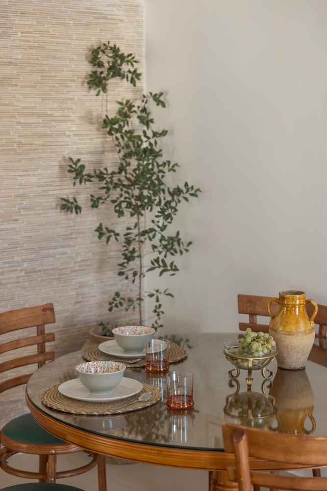 Dining still-life — glass table with wooden chairs, grapes on a footed dish, ceramic bowls, yellow amphora against the textured stone wall