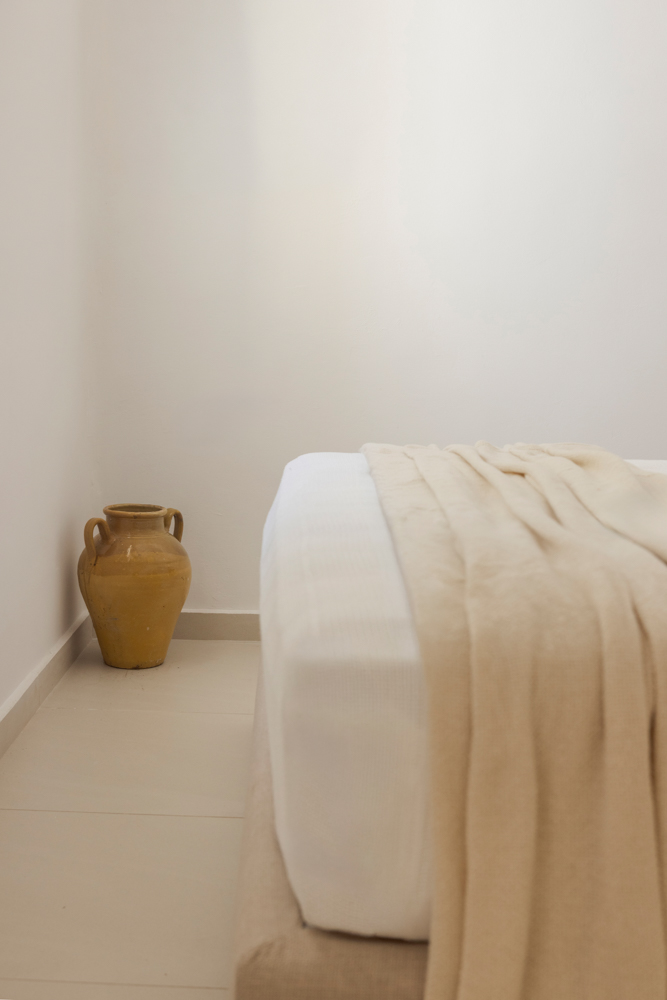 Detail: yellow amphora resting at the foot of a linen-clad bed