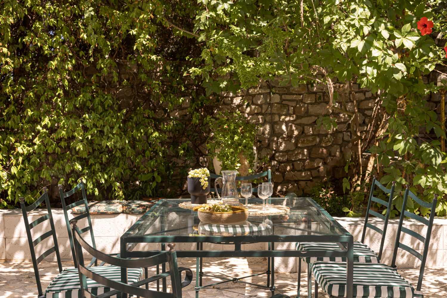Pergola dining table in the midday sun — grapes, jug of chilled wine, hibiscus blossoms overhead