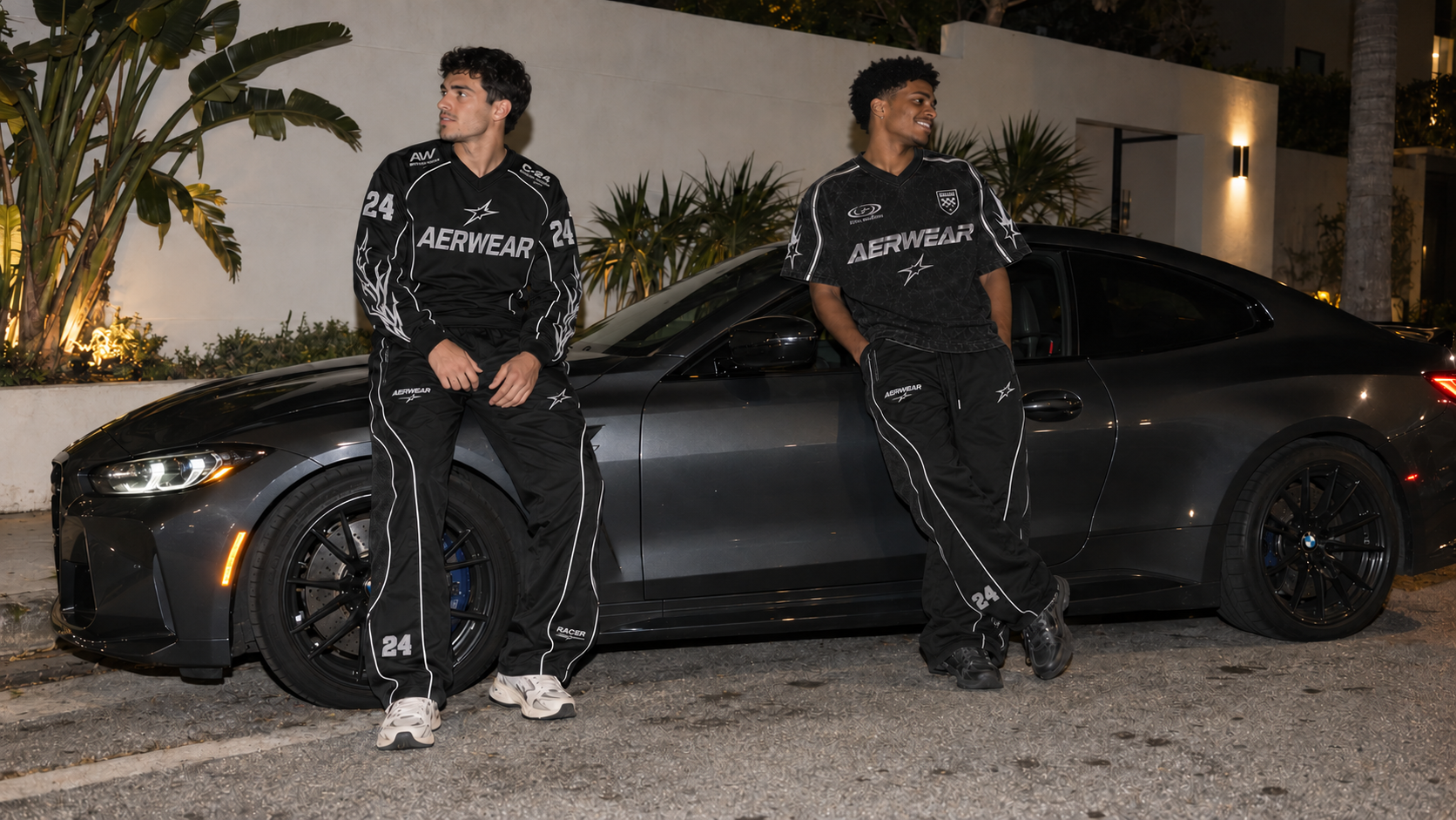 AERWEAR Racer Collection, two models in black racing jerseys and track pants beside a car at night.