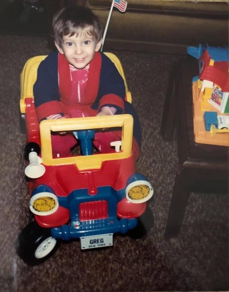 Toddler Greg in a red car with a 'GREG NEW YORK' plate